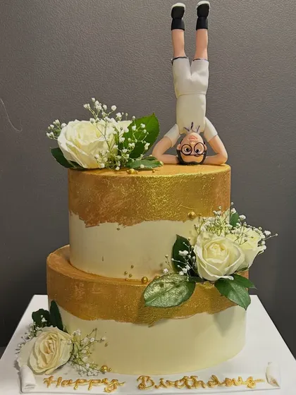 A fun and unique cake for a yoga lover! This two-tier cake features a hand-molded figure doing a headstand, surrounded by elegant white roses and gold accents.