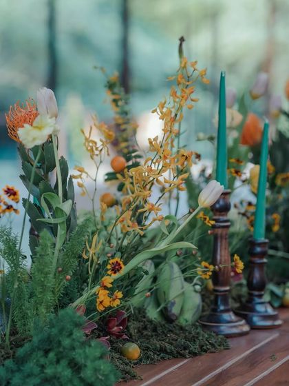 A long table on our outdoor deck is decorated with a lush, green and yellow floral runner. The arrangement includes tall green candles, creating a vibrant and natural centerpiece.