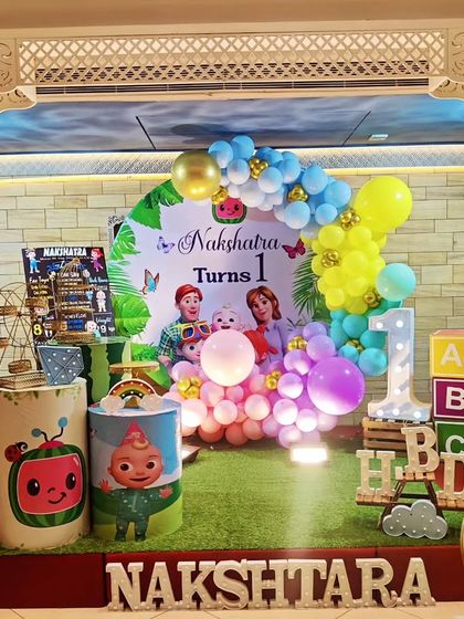A full stage setup for a Cocomelon-themed first birthday. The decor features a faux grass floor, the iconic yellow bus, and a 'NAKSHATARA' marquee.