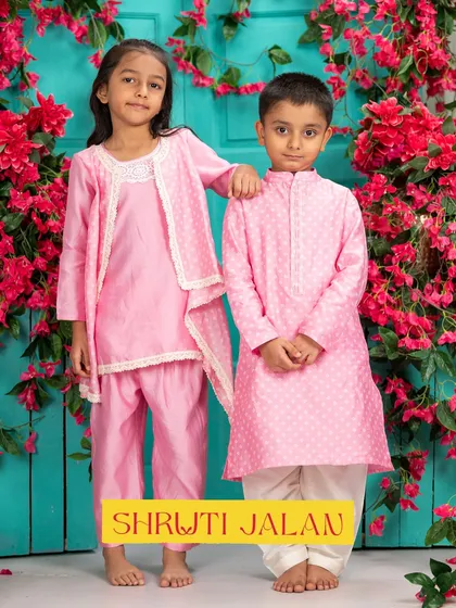 A brother and sister pose against a floral backdrop in their matching pink ensembles. Her draped kurta set with lace details is a perfect match for his dotted kurta.