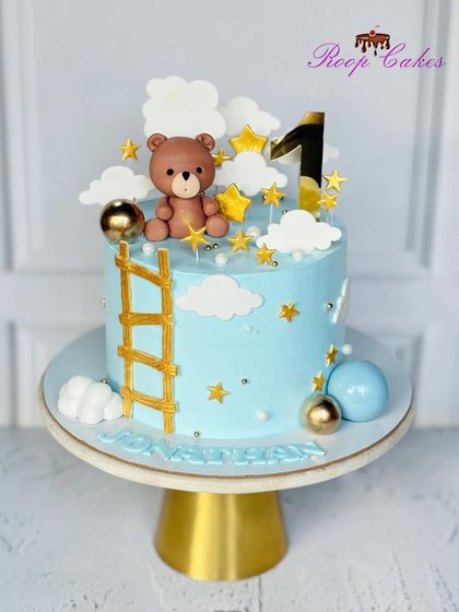 A sweet and dreamy cake for a first birthday. This design features a teddy bear climbing a golden ladder to the stars, surrounded by fluffy clouds and golden accents.