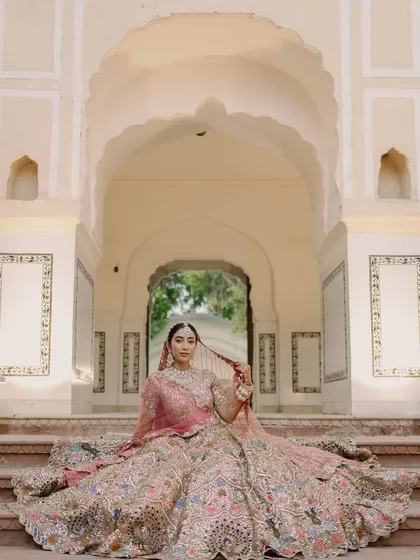 A regal bridal portrait showcasing the bride in her magnificent lehenga. Posed on the steps of a heritage property, this image highlights the intricate details of her attire and the elegance of the setting.