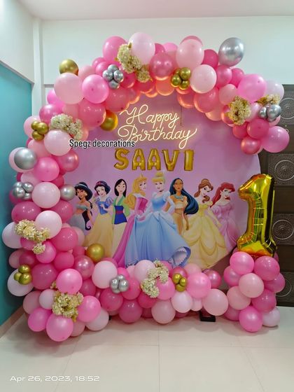 A Disney Princess dream-come-true for a first birthday. This grand setup includes a backdrop with all the iconic princesses, a pink balloon arch, and a gold foil number '1'.
