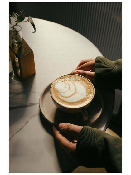 A lifestyle shot focusing on a pair of hands gently holding a cappuccino with beautiful latte art. The soft lighting and moody atmosphere create a cozy and inviting cafe scene.