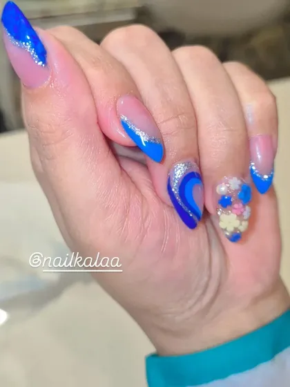 A close-up of a vibrant blue nail art design. This set features blue French tips, silver glitter lines, abstract swirls, and a stunning accent nail with 3D beaded flowers.