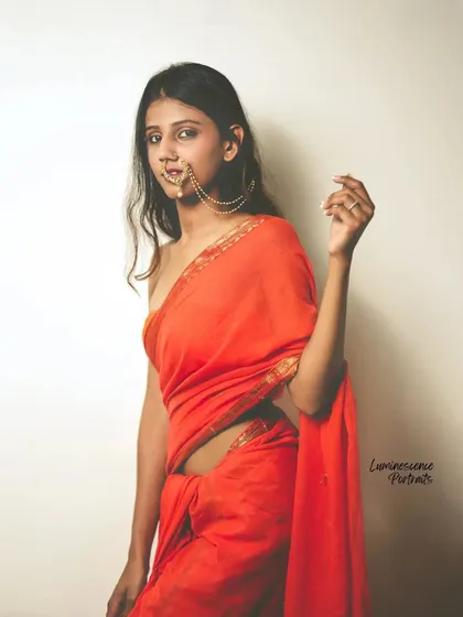 A confident and direct portrait of a model in a red saree and traditional nath, looking straight at the camera.