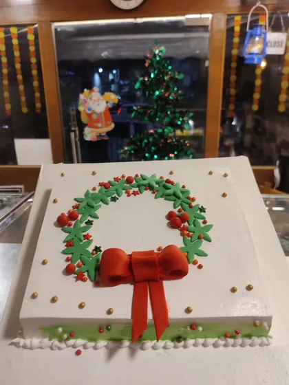Our Christmas wreath cake is an elegant choice for your holiday table. The simple square cake is topped with a beautifully piped green wreath, red berry details, and a festive red bow.