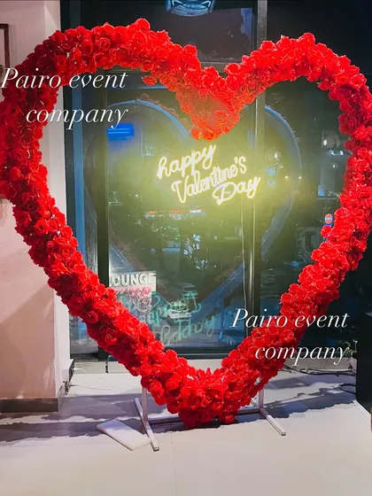 A bold Valentine's Day photo booth backdrop. The centerpiece is a large, free-standing heart frame made entirely of red roses, with a "Happy Valentine's Day" neon sign glowing from within.