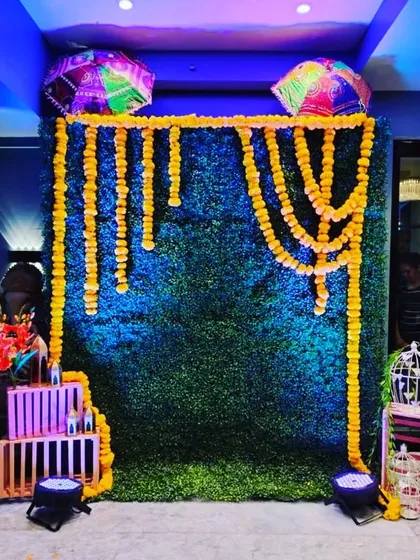 A simple yet beautiful Mehendi or Haldi corner. The setup uses a green grass wall decorated with marigold garlands and colorful Rajasthani umbrellas.