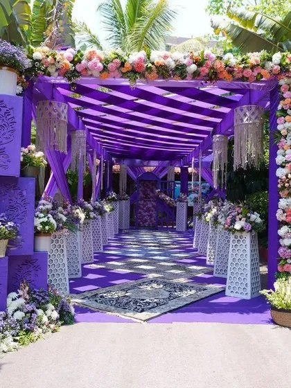 A bold and modern wedding entrance in a striking purple theme. The walkway is covered with a purple canopy and lined with decorative panels and floral arrangements, creating a unique and luxurious feel.