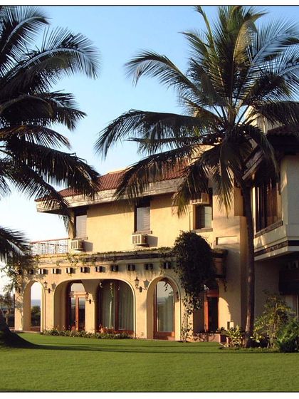 This sea-facing villa in Daman features traditional architectural elements like arched colonnades and a tiled roof, framed by beautiful palm trees that anchor it to its tropical location.
