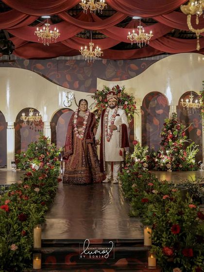 A couple stands proudly on their royal romance-themed wedding stage. The rich red roses, elegant draping, and chandeliers come together to create a picture of pure opulence.