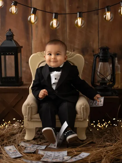 A little "Boss Baby" plays with prop money during his themed photoshoot. The rustic wooden background and string lights complete the look.