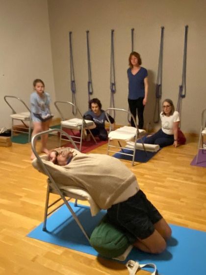 A class in Geneva, Switzerland, at Yoga-dea-Bains, where students are learning a supported restorative backbend using chairs and bolsters.