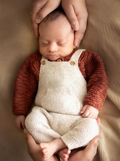 A "froggy" pose variation held safely by a parent's hands. This shot highlights the baby's tiny form and the protective embrace of his family.