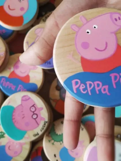 Holding up the main character herself. This Peppa Pig game piece is hand-painted on a smooth, round wooden tile, perfect for a game of memory.
