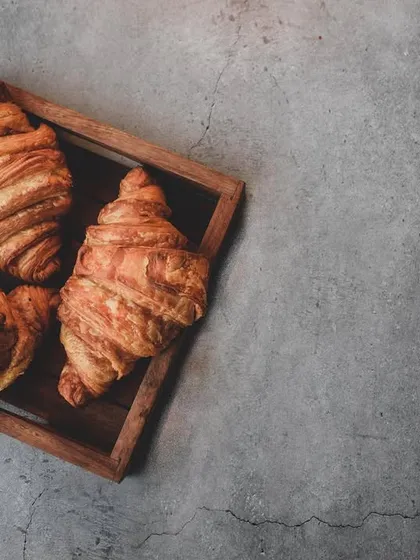 Flaky, buttery croissants captured in a moody, rustic style. The dark, textured background and top-down angle highlight the pastry's layers, creating an artisanal feel.