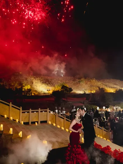 The couple shares a kiss as red fireworks explode in the sky above them, a perfectly timed moment of celebration and romance.
