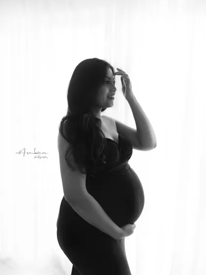 A soft and contemplative black and white portrait. The backlighting creates a beautiful rim light, separating her from the background and focusing on her gentle expression and the curve of her belly.
