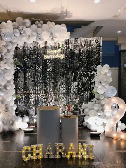 A stylish setup for a 21st birthday featuring a silver shimmer wall, a white and silver balloon arch, and large marquee numbers. The clean and modern look is perfect for milestone celebrations.