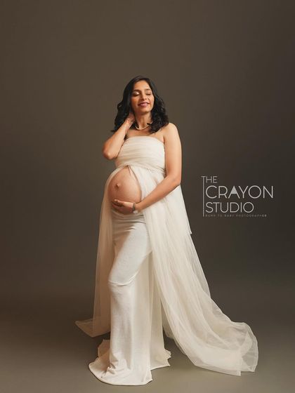 Dressed in ethereal white, she stands confidently, her bump beautifully defined. The simple grey backdrop and soft lighting create a serene and minimalist fine art portrait.
