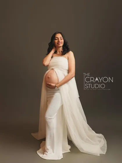 Dressed in ethereal white, she stands confidently, her bump beautifully defined. The simple grey backdrop and soft lighting create a serene and minimalist fine art portrait.