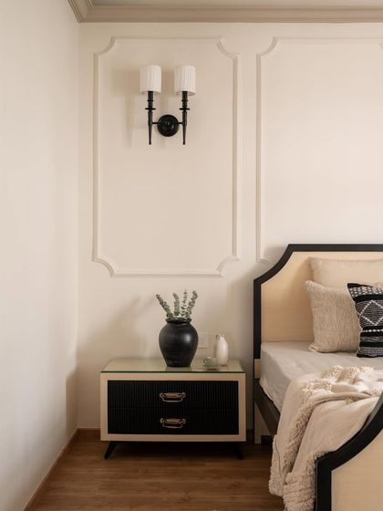 A classic black and cream bedroom from the Riviera Maison project. The bed features a gracefully curved headboard, flanked by a modern black nightstand and a traditional wall sconce.