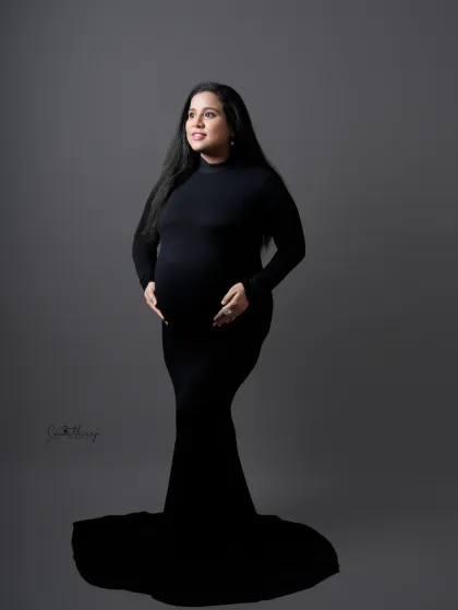 A classic full-length portrait of a mother-to-be in a black gown. The simple grey background and her graceful pose highlight her beautiful silhouette.