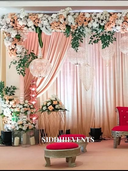 A beautiful stage setup where pastel pink curtains and hanging crystal chandeliers are complemented by a bold red chair and lush white and peach floral arrangements.