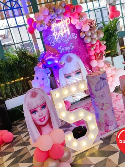 A vibrant Barbie-themed stage decoration for a 5th birthday. The setup includes a 'Happy Birthday' neon sign, marquee number, and Barbie character cutouts.