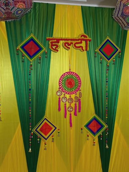 A simple and colorful Haldi backdrop with yellow and green drapes. The decor features a "Haldi" sign, a central dreamcatcher, and other traditional hangings with bells.