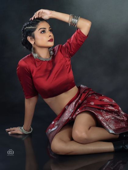 A seated portrait that showcases the modern draping of the red saree. The reflective floor and dark background create a polished, high-fashion studio look.