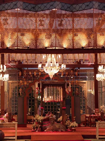 An evening view of a traditionally inspired venue space. The intricate wooden lattice work, grand chandeliers, and rich red carpet create a warm, opulent atmosphere that speaks of heritage and luxury.