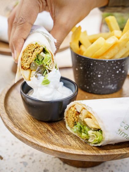 A Middle Eastern falafel wrap, cut in half and dipped in a creamy sauce. This action shot is dynamic and makes the food look fresh and delicious. The fries in the background complete the casual dining scene.