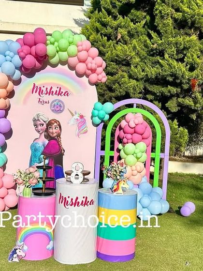A Frozen-themed party set in a lovely green garden. The decoration includes a pastel rainbow balloon arch, a backdrop with Elsa and Anna, and themed pedestals for the cake.