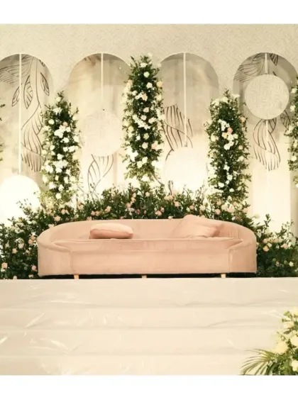 A clean and modern stage design using a white backdrop with minimalist line art. The decor is brought to life with vertical arrangements of white flowers and greenery, and a soft pink sofa adds a subtle touch of color.