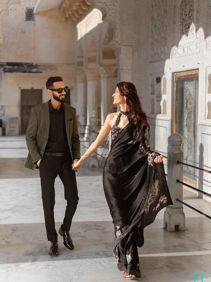 A candid walking shot through a palace corridor, capturing the effortless elegance of the couple. The movement in the bride's saree and their shared glance make this feel like a scene from a movie.