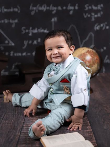A big, happy smile from a baby boy in his scholar-themed setup, sitting on the floor with a book.
