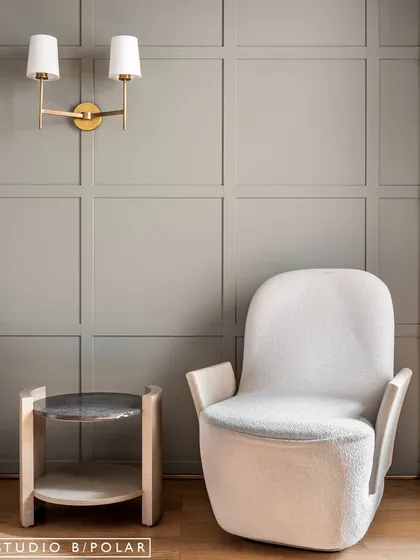 A cozy seating corner with a comfortable armchair and a small side table. The soft grey wall color and classic wall sconce create a serene and timeless feel.