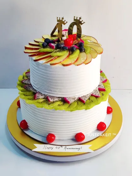 A two-tier mixed fruit anniversary cake. The cake is generously decorated with a variety of fresh, sliced fruits, making it a refreshing and beautiful centerpiece.