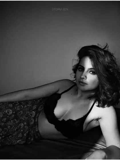 An intimate, moody portrait in black and white. The makeup is soft and natural, designed to look authentic in a bedroom setting with dramatic shadow play.