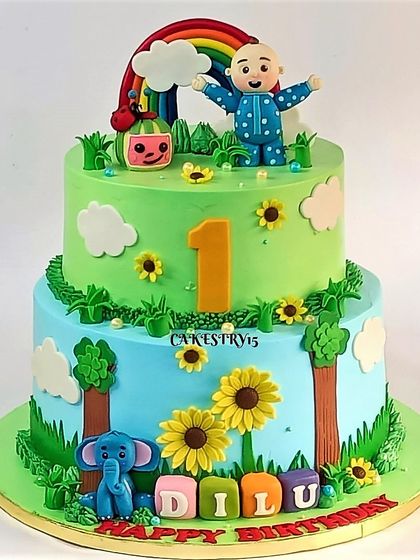 A charming two-tier Cocomelon cake with a nature theme. The green and blue tiers are decorated with fondant sunflowers, trees, an elephant, and the main Cocomelon characters.