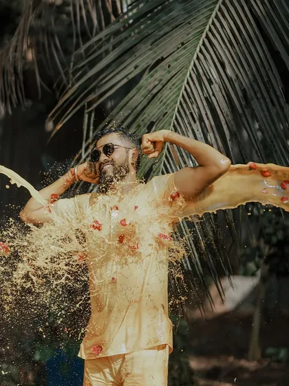 Capturing the pure, unadulterated fun of a Haldi ceremony. This high-energy shot freezes the moment the groom is joyfully drenched in turmeric paste and flower petals against a backdrop of lush palm leaves.
