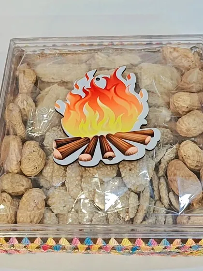 A simple and festive Lohri gift box with a clear lid and a bonfire sticker, filled with peanuts and sesame rewari.