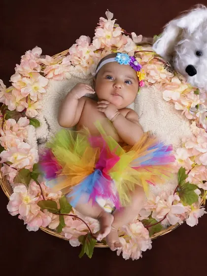 A colorful newborn photo with the baby wearing a rainbow tutu in a basket decorated with flowers. A soft toy dog adds a playful element to the scene.