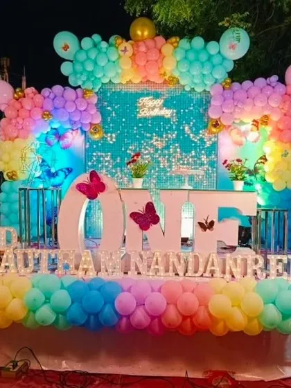 A colorful first birthday setup with a sequin backdrop, a rainbow of pastel balloons, and the baby's name in large light-up letters.
