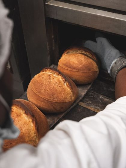 A baker carefully loads loaves of bread into the oven using a large wooden peel.