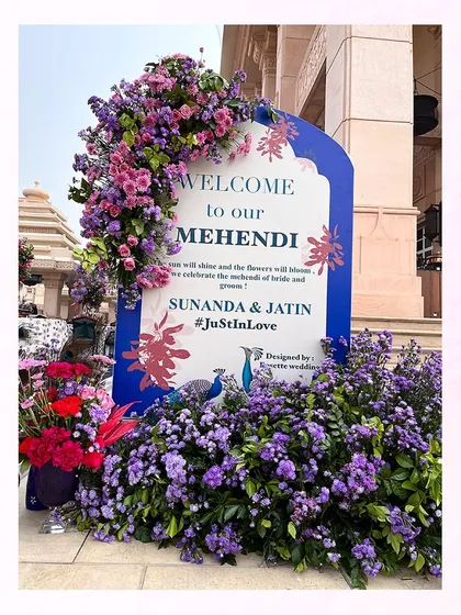 A welcome sign for the Mehendi, adorned with a cascade of purple and pink flowers, tying into the peacock theme.