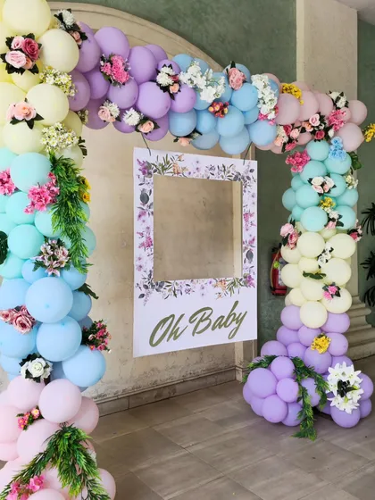 A beautiful "Oh Baby" photo frame surrounded by a pastel rainbow balloon arch. The arch is decorated with delicate flowers, making it perfect for a spring or garden-themed baby shower.