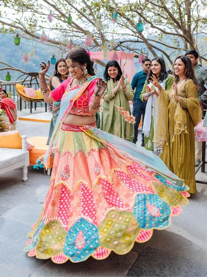 A bride twirls in her vibrant, multi-colored lehenga, her joy evident as her friends clap for her.